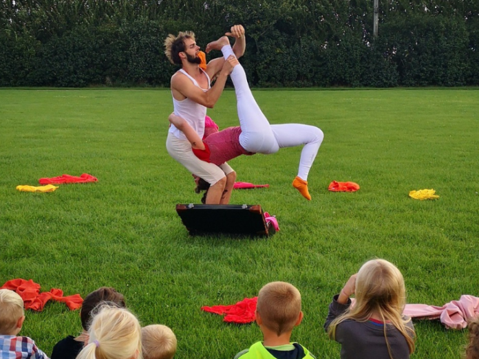 En kvinde og en mand i gymnastikdragter laver akrobatik på en græsplæne, mens børn kigger på